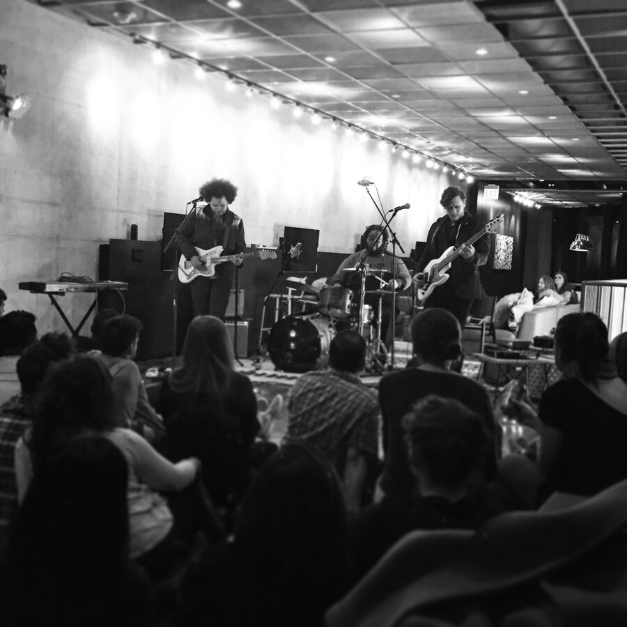 Black and white photo of Hunter-gatherer playing in a concrete room under a line of string lights to a seated crowd