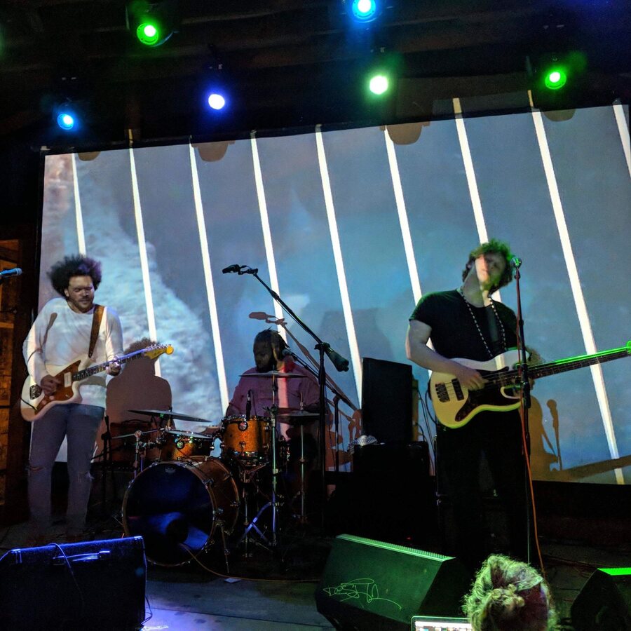 Hunter-gatherer in front of a striped projection screen on a lit club stage, the singer's hair mid-whip, drummer and bassist either side